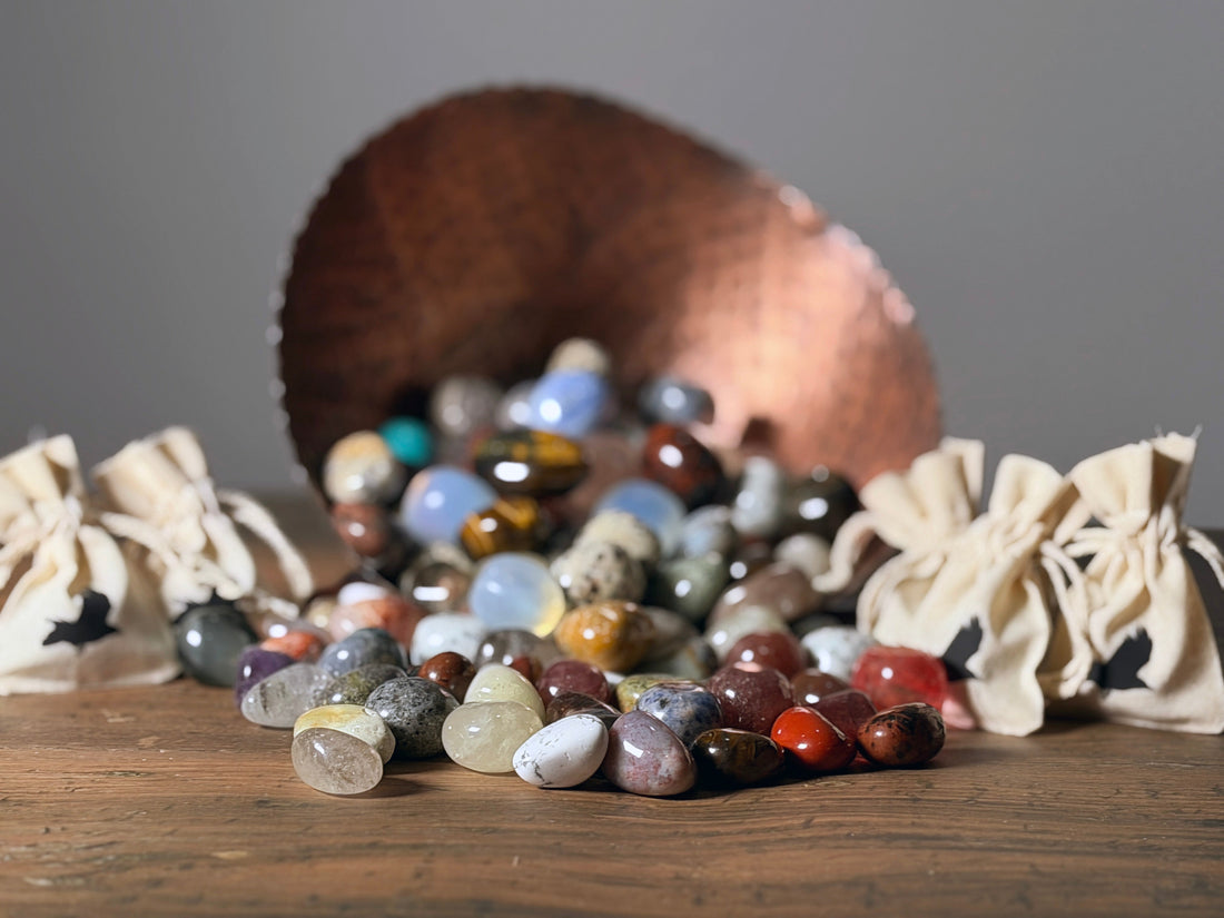 A hammered copper bowl filled with polished stones surrounded by natural cotton drawstring bags stamped with the Copper Crow Fiber Co. crow logo, each containing a polished stone.